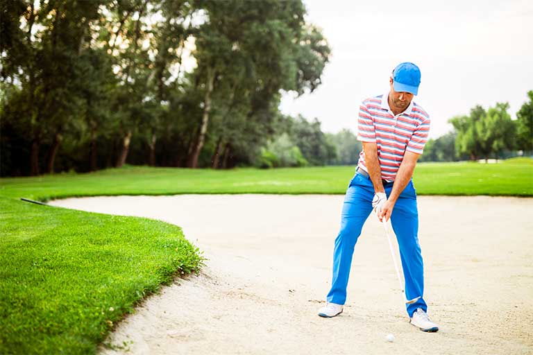 How to Hit a Bunker Shot 1 A golfer in a blue and white outfit prepares to hit a ball out of a sand bunker on a green golf course. Trees line the background. This is how to hit a bunker shot.