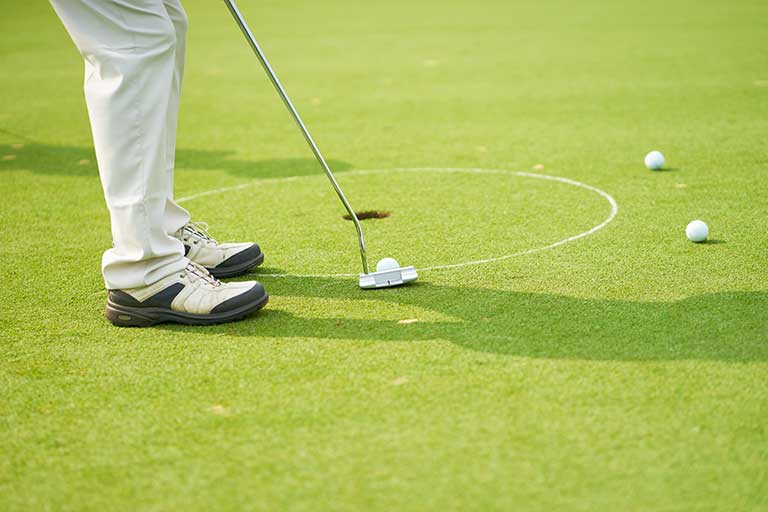 10 Golf Practice Drills to Lower Your Scores 1 A man is putting a golf ball into a hole while practicing on how to lower his golf score.