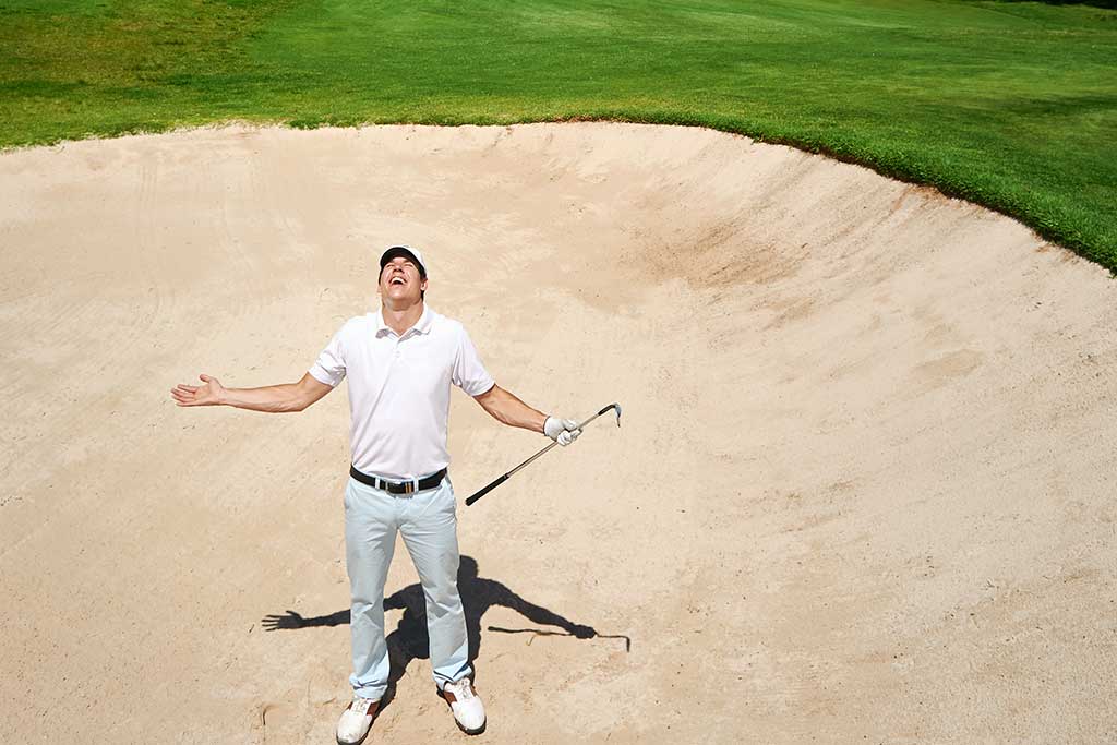 How to Handle Conflict on the Golf Course 1 A frustrated golfer in white attire stands in a sand bunker, looking upwards with his arms spread out and holding a golf club.