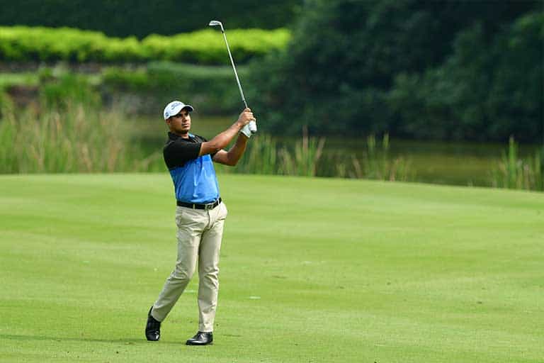 How to Hit a 50-Yard Pitch Shot 1 A golfer mid-swing on a green golf course, wearing a blue and black shirt, beige pants, and a white cap. Trees and bushes are visible in the background. This is how to hit a 50 yard pitch shot.