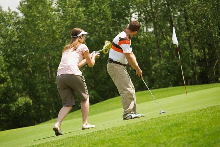 Golf Etiquette: 10 Encouraged Rules of the Game 1 Two golfers on the putting green, observing golf etiquette, man taking a shot while woman tries to distract him.