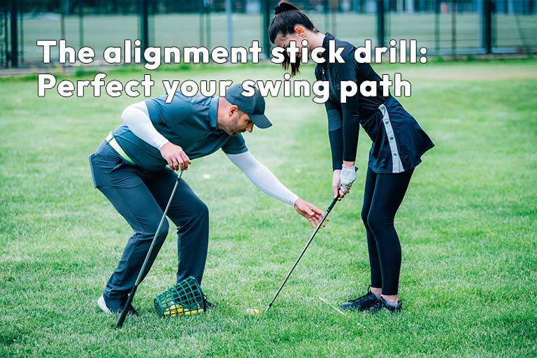 A golf instructor helps a golfer with her swing alignment using alignment sticks on a grassy area. The text on the image reads, "The alignment stick drill: Perfect your swing path.