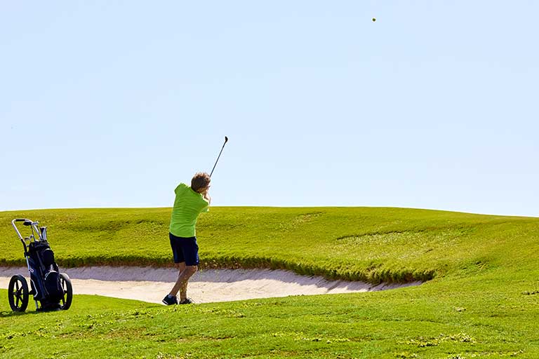 How to Hit a Golf Ball from an Uneven Lie 1 Golfer in green shirt swinging at a ball on a grassy course by a sand bunker, with a golf bag on a trolley nearby. The golfer needs to Hit a Golf Ball from an Uneven Lie