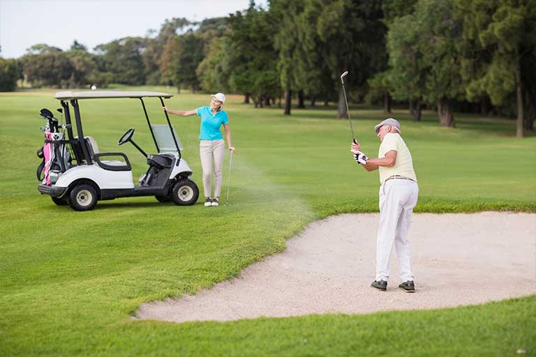Bunker consistency: The dollar bill drill for sand shots 1 An older man plays a sand shot on a golf course while an older woman stands by a golf cart, waving, surrounded by trees.