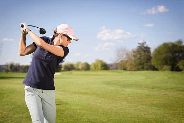 How to Hit a Golf Ball: A Beginner’s Guide 1 A woman in a pink cap and sunglasses swings a golf club on a sunny, grassy course.