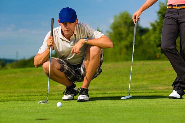 How to read greens: 5 simple steps to improve your putting accuracy 1 A man crouching to line up a putt on a sunny golf course, with another player standing nearby.