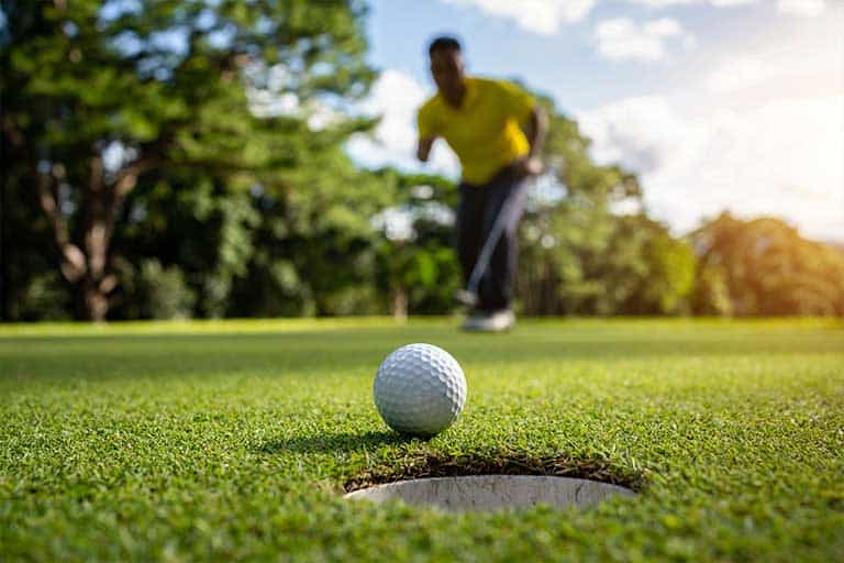 10 tips to improve your short game 1 A man is practicing his short game by sinking a golf ball into a hole.
