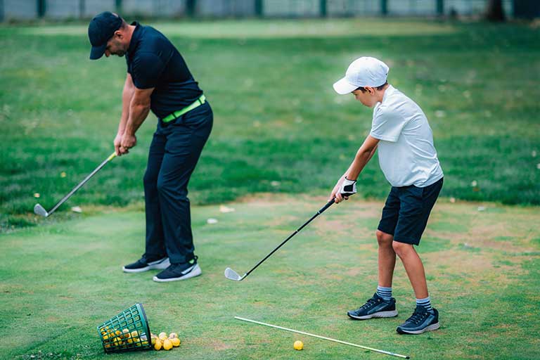 The alignment stick drill: Perfect your swing path 1 An adult and a child practice golf swings on a grassy area. The child wears a white cap and holds a golf club. A basket of yellow golf balls is on the ground near them.