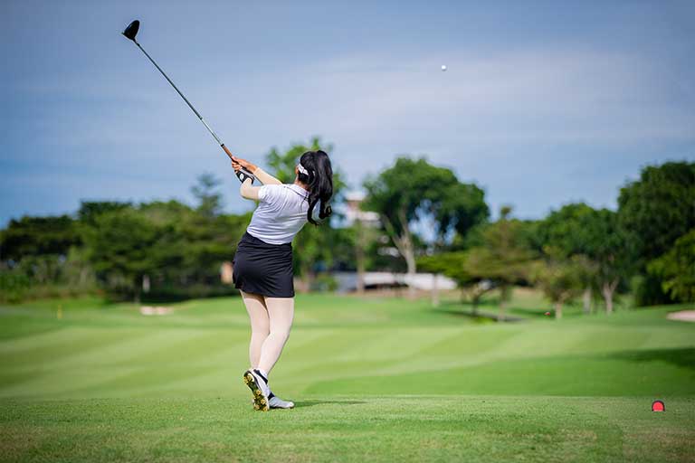 Tempo training: The metronome drill for a smooth swing 1 A person in a white shirt and black shorts swings a golf club on a green golf course with trees and a bright sky in the background.